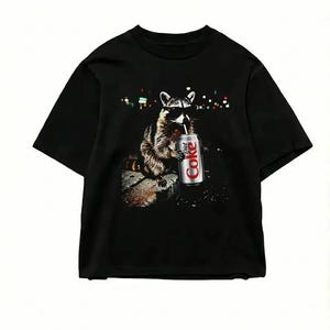 May include: Black t-shirt featuring a raccoon wearing sunglasses and drinking from a Diet Coke can. The raccoon is depicted in a detailed illustration, set against a blurred background of city lights. The shirt is a casual wear item.