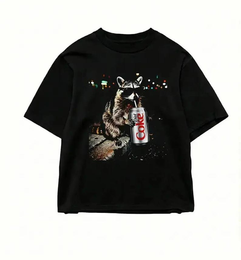 May include: Black t-shirt featuring a raccoon wearing sunglasses and drinking from a Diet Coke can. The raccoon is depicted in a detailed illustration, set against a blurred background of city lights. The shirt is a casual wear item.