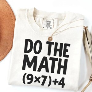 May include: Cream-colored t-shirt with the phrase "DO THE MATH" in bold black letters, followed by the equation (9x7)+4. A gold necklace with a pendant rests on the shirt. A brown hat is partially visible in the upper left corner.