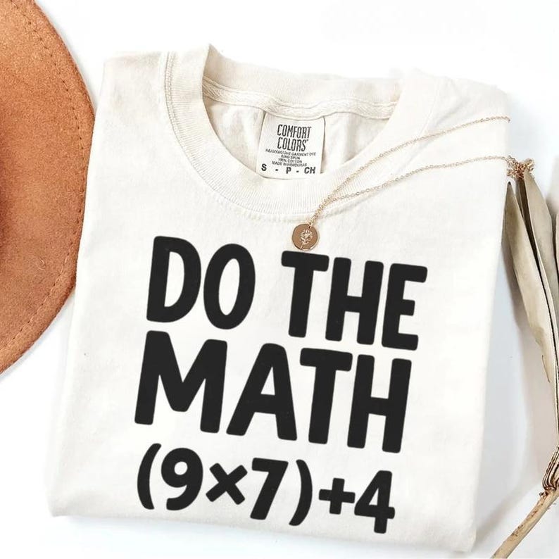 May include: Cream-colored t-shirt with the phrase "DO THE MATH" in bold black letters, followed by the equation (9x7)+4. A gold necklace with a pendant rests on the shirt. A brown hat is partially visible in the upper left corner.