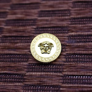 Puede incluir: Un botón circular de color dorado con fondo blanco y un diseño de cabeza de Medusa de color dorado. El botón tiene un borde decorativo.