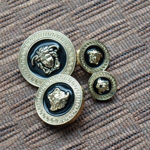 May include: Four gold-colored buttons with black enamel and a raised gold design of a face. The buttons are arranged in a semi-circle on a brown textured surface.