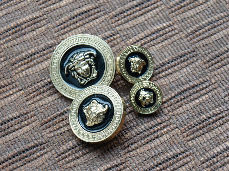 May include: Four gold-colored buttons with black enamel and a raised gold design of a face. The buttons are arranged in a semi-circle on a brown textured surface.