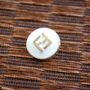 Puede incluir: Un pin redondo blanco y dorado con las letras "FF" doradas. El pin está sobre un fondo tejido marrón. Probablemente sea un accesorio de moda.