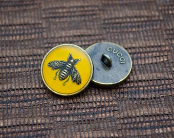May include: Two gold-toned buttons, one with a yellow background and a black bee design, and the other with the word "GUCCI" engraved on it.