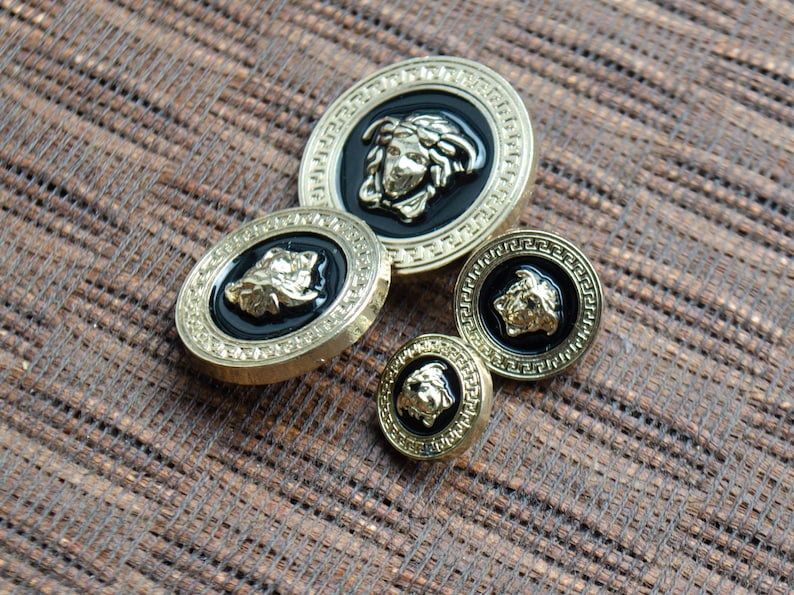 May include: Four gold-toned buttons with black enamel and raised lion head designs. The buttons are different sizes and have a decorative border.
