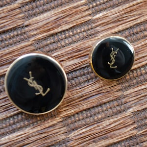 May include: A pair of black and gold round earrings. Each earring has a black centre with a gold logo and a gold border. The earrings are presented on a textured brown surface.