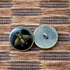Puede incluir: Dos botones redondos de color dorado. Un botón tiene un fondo negro con un diseño de abeja dorada. El otro botón tiene la palabra "GUCCI" grabada.