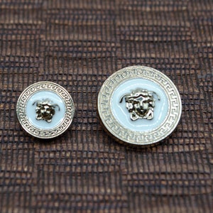 May include: Two gold-toned buttons with a white enamel background and a gold-toned Medusa head design. The buttons have a decorative border.