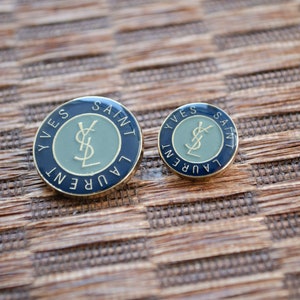 Puede incluir: Dos pines de metal dorado con el logotipo de Yves Saint Laurent. El pin más grande tiene un anillo exterior azul y un círculo interior verde con el logotipo en oro. El pin más pequeño tiene un anillo exterior dorado y un círculo interior verde con el logotipo en oro.