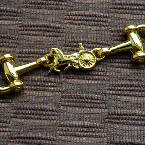 Könnte beinhalten: Goldfarbener Metall-Pferdekutschen-Anhänger mit zwei goldfarbenen Metall-Gebiss-Verschlüssen. Der Anhänger ist mit einer goldfarbenen Metallkette an den Verschlüssen befestigt.