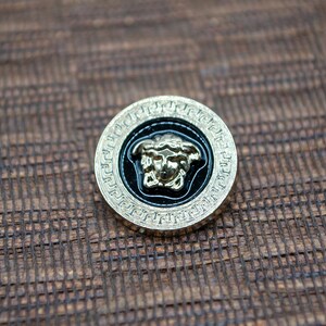 May include: A gold-colored button with a black background and a gold-colored Medusa head design. The button has a decorative border.