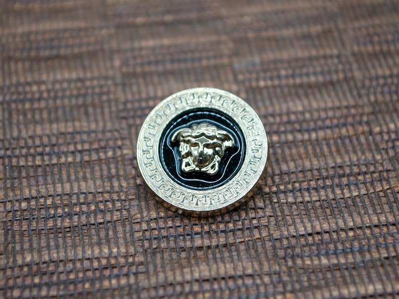 May include: A gold-colored button with a black background and a gold-colored Medusa head design. The button has a decorative border.