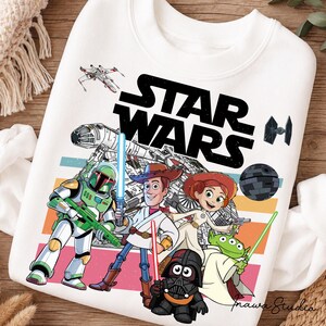 Op de afbeelding: Een wit sweatshirt met een grafische print van Star Wars-personages die als speelgoed zijn afgebeeld. Het ontwerp bevat Boba Fett, Woody, Jessie en een alien met lichtzwaarden, samen met de Millennium Falcon, X-wing, TIE fighter en Death Star.