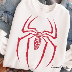 May include: A white crewneck sweatshirt featuring a large red spider graphic on the front. The body of the spider is composed of the text "WITH GREAT POWER COMES GREAT RESPONSIBILITY" in a bold, stylized font. The design is centered on the chest.
