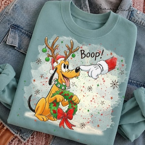 May include: A light blue crewneck sweatshirt featuring a graphic of a yellow dog wearing reindeer antlers and a Santa hat, holding a green wreath with a red bow. A gloved finger touches the dog's nose with the text Boop! above the dog.