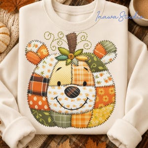 May include: A cream-coloured crewneck sweatshirt featuring a large, central graphic of a bear face constructed from various patchwork fabric patterns in shades of orange, yellow, green, and brown. The design includes a button nose and stitched details.