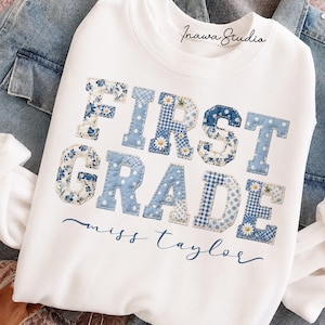 Könnte beinhalten: Ein weißes Sweatshirt mit Rundhalsausschnitt und dem Schriftzug FIRST GRADE in großen, blau-weiß gemusterten Patchwork-Buchstaben mit Blumen- und Vichy-Karomustern. Darunter steht der Name miss taylor in dunkelblauer Schreibschrift.