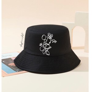 May include: Black bucket hat with a white outline drawing of a cartoon character holding a balloon. The character is wearing a dress and has a bow on its head.