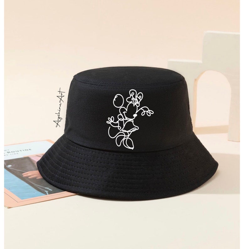 May include: Black bucket hat with a white outline drawing of a cartoon character holding a balloon. The character is wearing a dress and has a bow on its head.