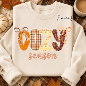 Peut inclure: Un sweat-shirt à col rond couleur crème portant l'inscription "COZY season" dans une police décorative. Les lettres de "COZY" sont remplies de pois orange, de carreaux, de spirales jaunes et de feuilles d'automne brunes, avec deux nœuds.
