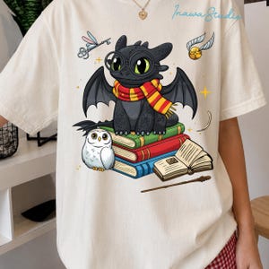 Może przedstawiać: Kremowy t-shirt z grafiką czarnego smoka w okrągłych okularach i czerwono-żółtym pasiastym szaliku, siedzącego na stosie książek obok białej sowy, latającego klucza, złotego znicza, otwartej książki i różdżki.