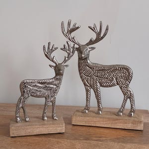 May include: Two silver deer ornaments with detailed antlers and textured bodies. Each deer is mounted on a rectangular wooden base. The ornaments are displayed on a wooden surface against a neutral background.