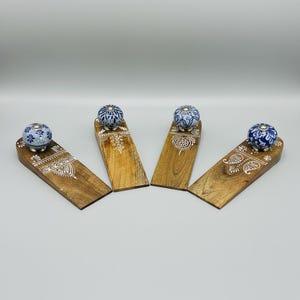 May include: Four wooden doorstops with decorative ceramic knobs. The doorstops are made of light brown wood with white painted designs. The ceramic knobs are blue and white with floral patterns. The doorstops are arranged on a white surface.