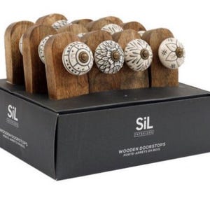May include: A set of wooden doorstops with ceramic knobs. The knobs have a white background with black geometric designs. The doorstops are packaged in a black box with the text "SIL INTERIORS WOODEN DOORSTOPS PORTE-ARRETS EN BOIS" printed on the front.