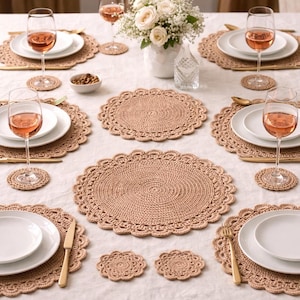 Puede incluir: Una mesa puesta con manteles individuales y posavasos de ganchillo beige, y platos blancos. Cubiertos dorados y copas de vino llenas de vino rosado se disponen alrededor de un jarrón con flores blancas.