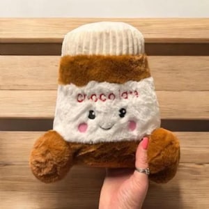 May include: A soft toy resembling a jar of chocolate spread, with a white top, brown body, and brown arms. The word "chocola" is embroidered in red. The toy has a smiling face with pink cheeks.