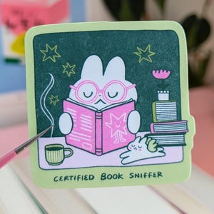 May include: A square sticker featuring a cartoon bunny with glasses, reading a pink book, and the text "CERTIFIED BOOK SNIFFER". The sticker is placed on a stack of books with colourful spines.
