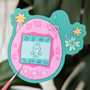 Könnte beinhalten: Ein pinkfarbener und türkisfarbener Aufkleber mit einem Cartoon-Tamagotchi-Design. Der Bildschirm des digitalen Haustiers zeigt einen Hasencharakter. Der Aufkleber ist mit Stern- und Planetenakzenten sowie einer türkisfarbenen Kette mit Stern- und Blumenanhängern verziert.