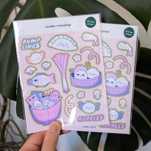 Puede incluir: Dos hojas de pegatinas con ilustraciones de dibujos animados de alb&oacute;ndigas y fideos en rosa, azul y blanco. Las pegatinas tienen el texto "noodles + dumplings", "DUMP LINGS" y "NOODLES". El fondo es una cuadr&iacute;cula rosa claro.