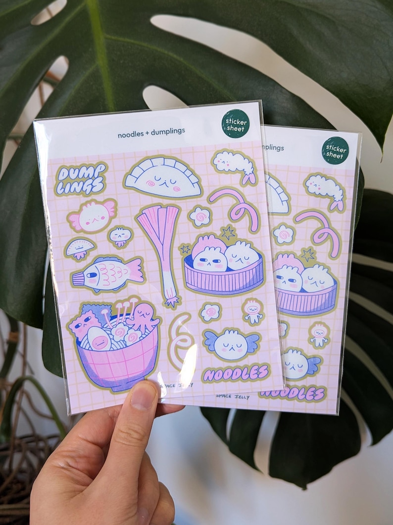 Puede incluir: Dos hojas de pegatinas con ilustraciones de dibujos animados de alb&oacute;ndigas y fideos en rosa, azul y blanco. Las pegatinas tienen el texto "noodles + dumplings", "DUMP LINGS" y "NOODLES". El fondo es una cuadr&iacute;cula rosa claro.