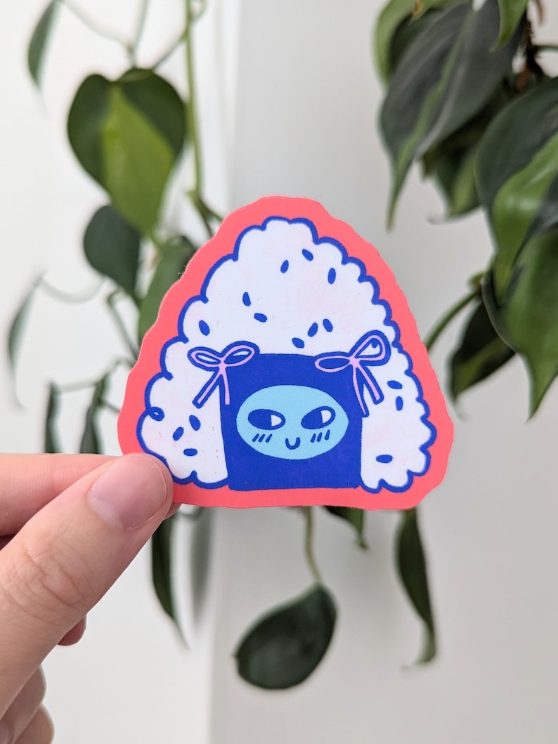 May include: A pink and blue sticker of a cartoon onigiri, a Japanese rice ball, with a smiling face and a blue headband. The onigiri is surrounded by white rice grains.