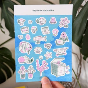 May include: A sheet of stickers with an ocean theme. The stickers feature various sea creatures and office-related items, such as fish, octopuses, and a printer. The stickers are in shades of pink, blue, and white, with the text "days at the ocean office".