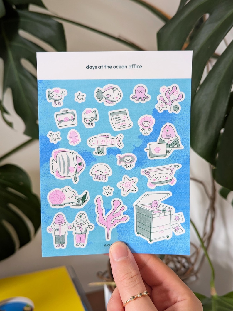 May include: A sheet of stickers with an ocean theme. The stickers feature various sea creatures and office-related items, such as fish, octopuses, and a printer. The stickers are in shades of pink, blue, and white, with the text "days at the ocean office".