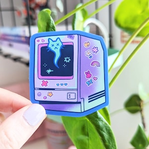 Puede incluir: Una pegatina con un diseño de ordenador retro y un borde azul. La pantalla del ordenador muestra un gato fantasma y estrellas. Gráficos adicionales incluyen corazones, flores, un arco iris y una mariposa. La pegatina está sujeta por una mano.