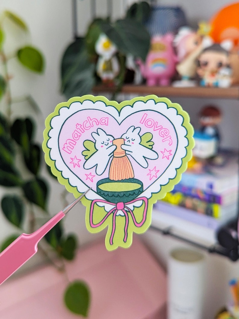 Puede incluir: Una pegatina en forma de coraz&oacute;n con fondo rosa claro y borde verde festoneado. La pegatina presenta dos conejitos de dibujos animados con alas, un cuenco de matcha y las palabras "matcha lover". Estrellas rosas y un lazo verde completan el dise&ntilde;o.