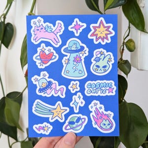 May include: A blue sticker sheet featuring various cosmic-themed illustrations. Designs include a pink cat with wings, a heart, stars, a UFO, and a green alien. The text "Cosmic Cutie" is also present.