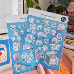May include: Two sticker sheets with a blue background and various pink and white sea creature illustrations. The text "days at the ocean office" is printed at the top. The stickers are in a clear plastic sleeve.