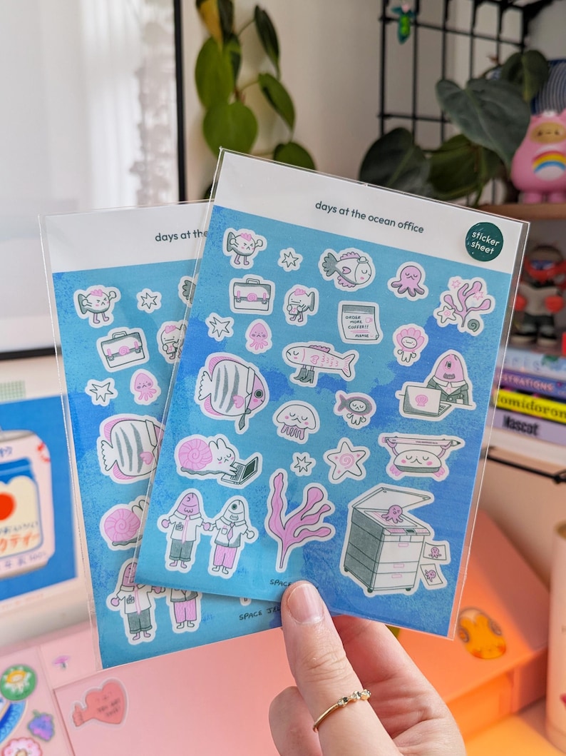 May include: Two sticker sheets with a blue background and various pink and white sea creature illustrations. The text "days at the ocean office" is printed at the top. The stickers are in a clear plastic sleeve.