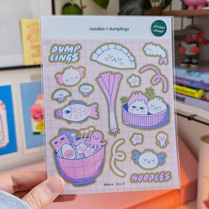 Puede incluir: Una l&aacute;mina de pegatinas de pl&aacute;stico transparente con ilustraciones coloridas de fideos y empanadillas. Las pegatinas incluyen im&aacute;genes de un pez, un cuenco de ramen y varios dise&ntilde;os de empanadillas. El texto en la l&aacute;mina dice "noodles + dumplings", "DUMP LINGS", "NOODLES" y "SPACE JELLY".