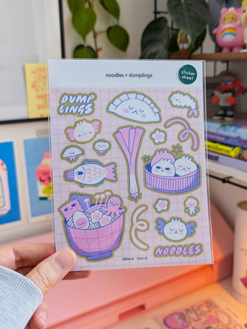 Puede incluir: Una l&aacute;mina de pegatinas de pl&aacute;stico transparente con ilustraciones coloridas de fideos y empanadillas. Las pegatinas incluyen im&aacute;genes de un pez, un cuenco de ramen y varios dise&ntilde;os de empanadillas. El texto en la l&aacute;mina dice "noodles + dumplings", "DUMP LINGS", "NOODLES" y "SPACE JELLY".
