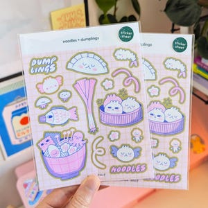 Puede incluir: Dos hojas de pegatinas con ilustraciones de fideos y empanadillas. Las pegatinas son rosas y doradas, con el texto "noodles + dumplings", "DUMP LINGS" y "NOODLES". Las pegatinas est&aacute;n empaquetadas en una funda de pl&aacute;stico transparente.