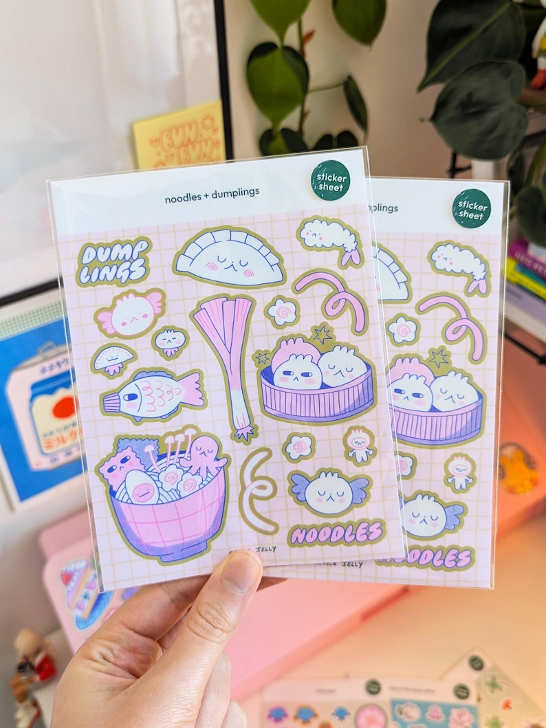 Puede incluir: Dos hojas de pegatinas con ilustraciones de fideos y empanadillas. Las pegatinas son rosas y doradas, con el texto "noodles + dumplings", "DUMP LINGS" y "NOODLES". Las pegatinas est&aacute;n empaquetadas en una funda de pl&aacute;stico transparente.