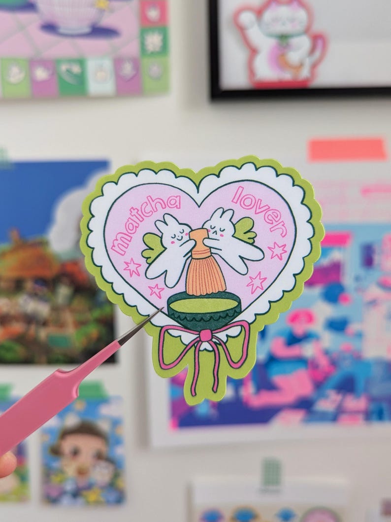 Puede incluir: Una pegatina en forma de coraz&oacute;n con las palabras "matcha lover" en letras rosas. La pegatina presenta dos conejitos de dibujos animados con alas, un cuenco de matcha y un batidor. La pegatina tiene un borde festoneado verde y un lazo verde en la parte inferior.