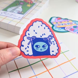May include: A red and white sticker with a blue and white cartoon face inside. The cartoon face has a happy expression with two large eyes and a small mouth. The sticker is shaped like a triangle and has a white border.