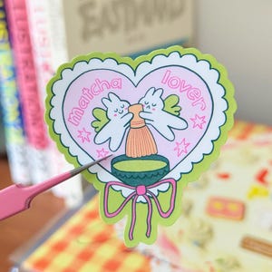 Puede incluir: Una pegatina en forma de coraz&oacute;n con un borde festoneado verde y un interior rosa. La pegatina presenta dos conejitos de dibujos animados con alas, un batidor de matcha y las palabras "matcha lover". Una cinta verde en la parte inferior.